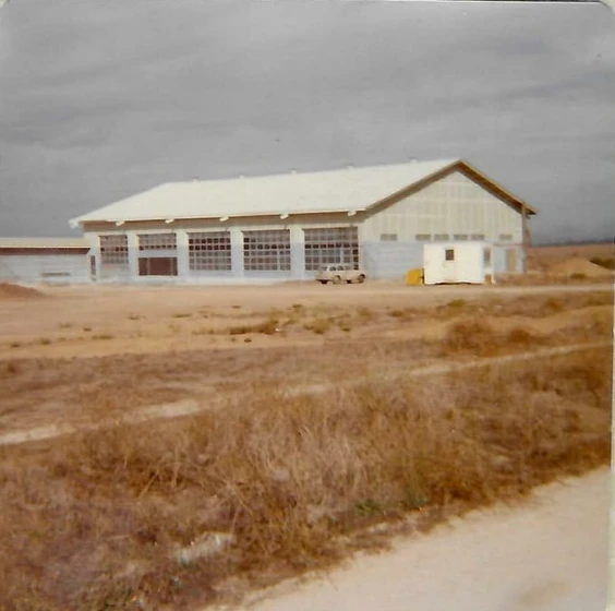 An old, large building with multiple garage doors, set in a barren field with overcast skies.