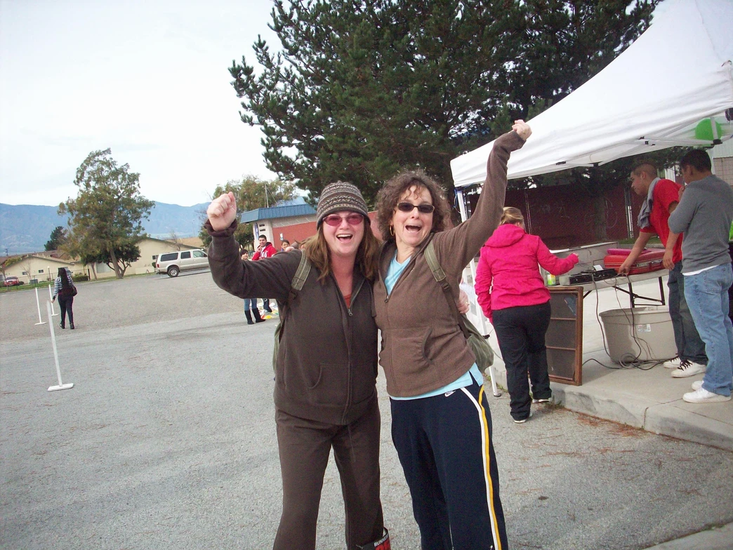 Two joyous women raising their arms in celebration, with a background of an outdoor event including a tent and other people.