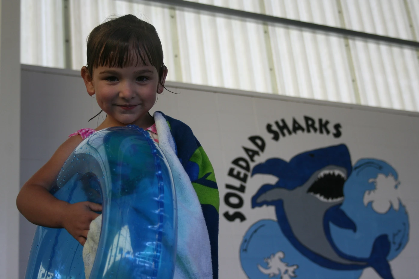 A smiling child with a towel and inflatable ring by a "Soledad Sharks" sign.