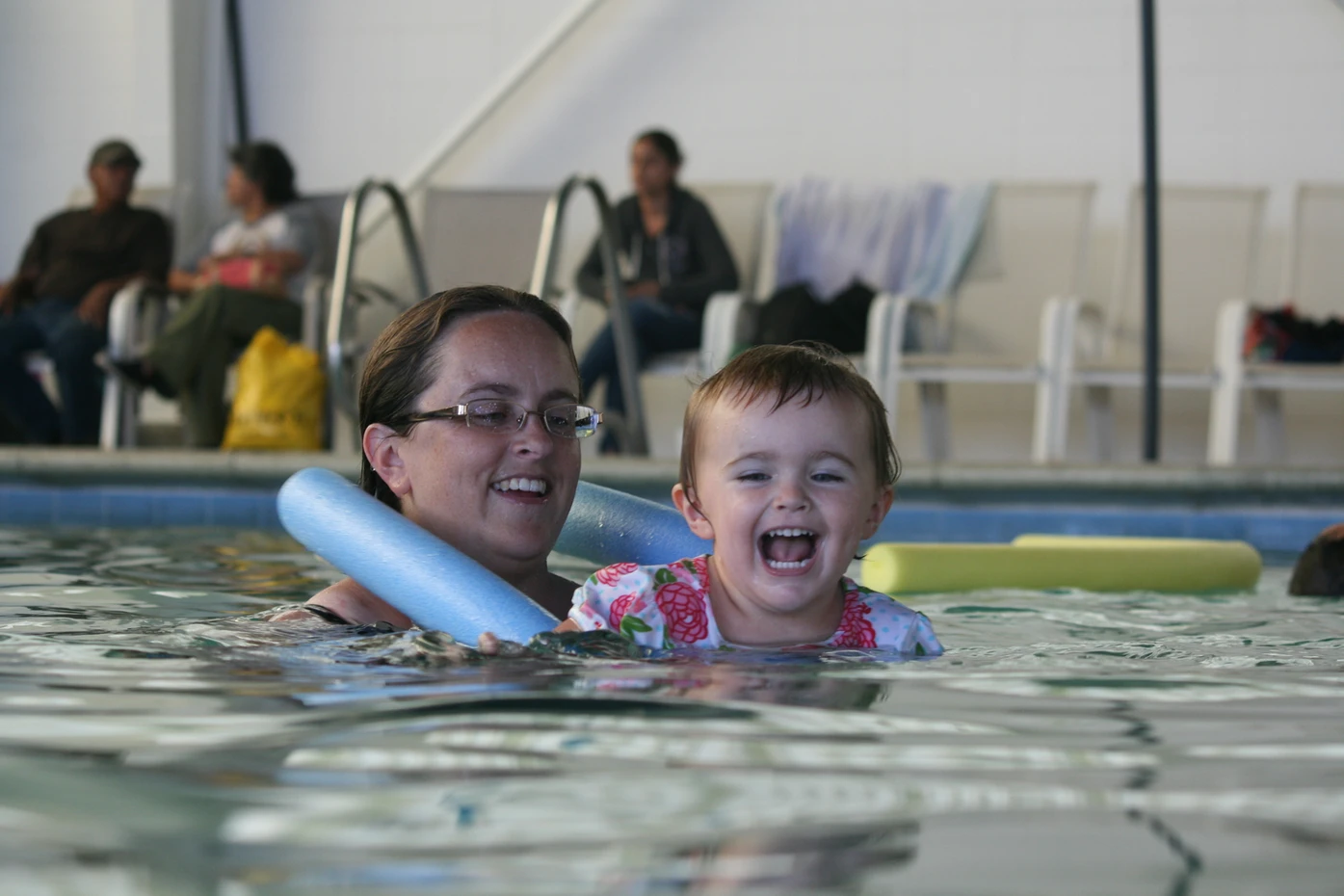 An adult and a child are enjoying the pool, surrounded by floating toys and people sitting poolside. They seem happy.