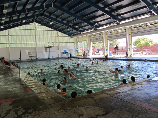 Indoor swimming pool with people swimming and socializing, partial roof cover, and daylight.