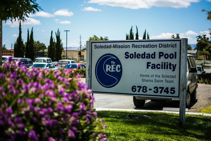 Sign for Soledad Pool Facility with contact number, flowers in foreground, cars and trees in background.