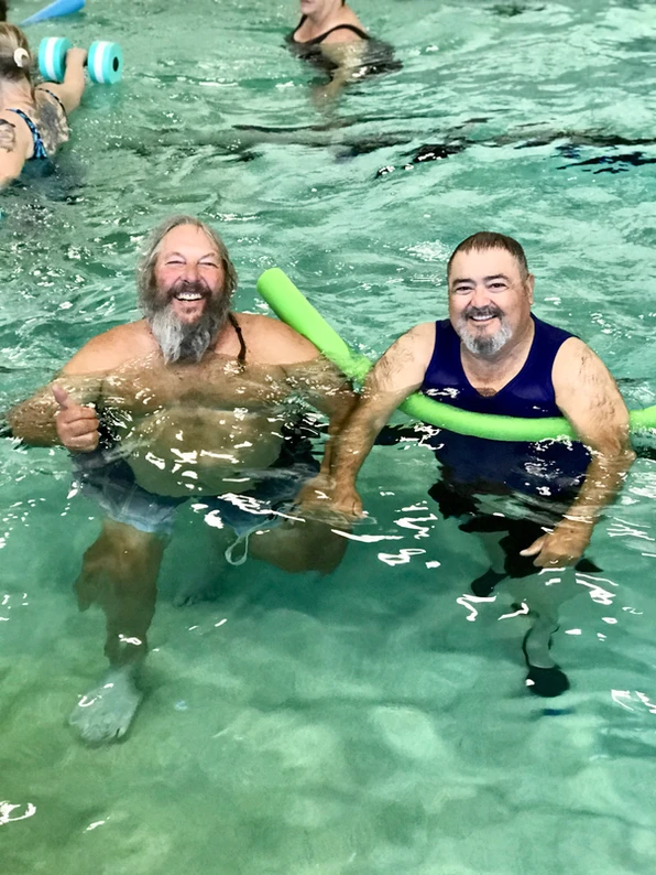 Two people smiling in a pool, one with a green noodle float.