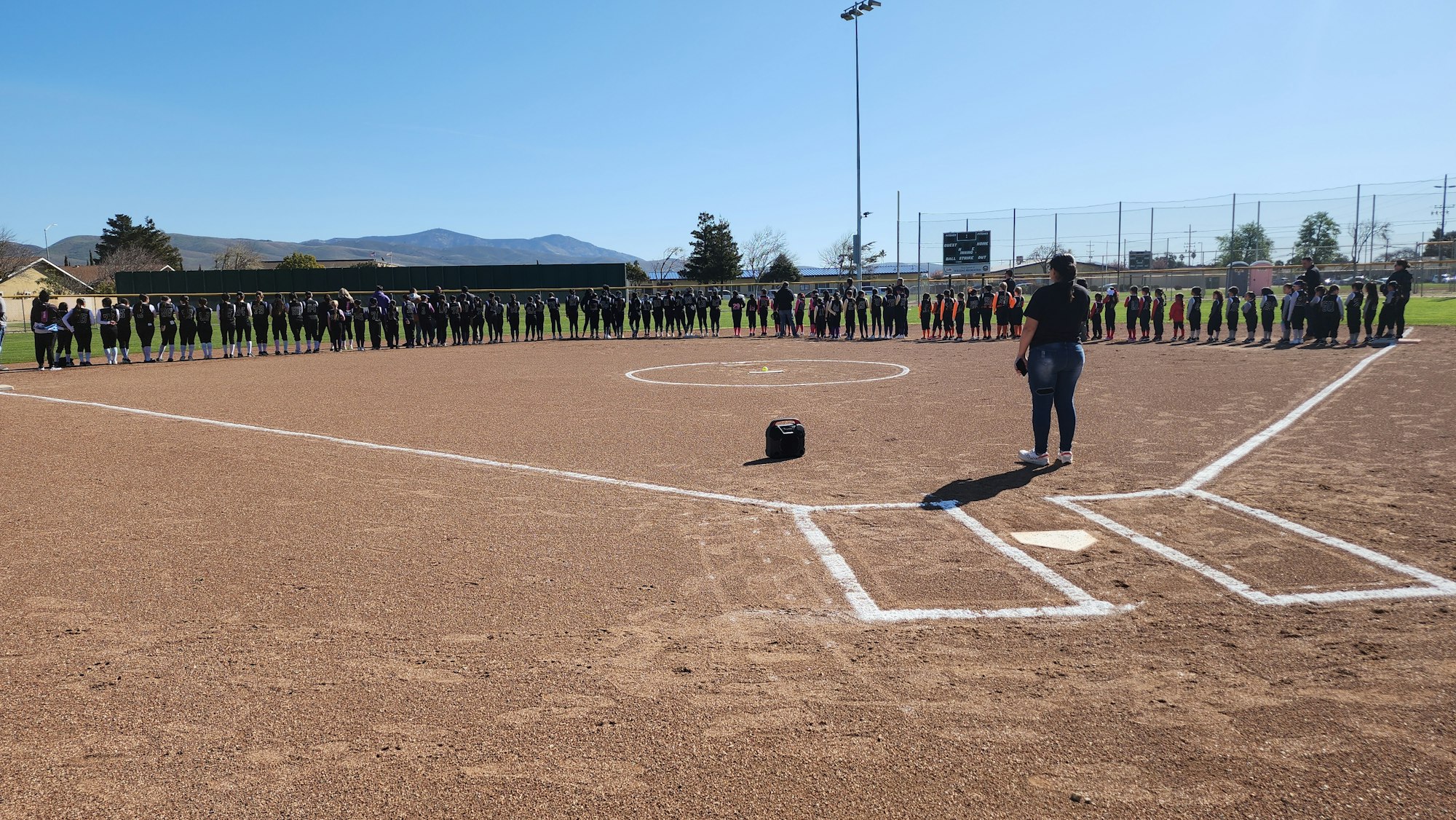 People are lined up along the softball field baseline, possibly for a ceremony or event.