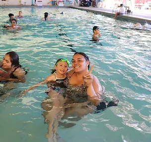 A child and an adult smiling in a swimming pool, surrounded by other swimmers.