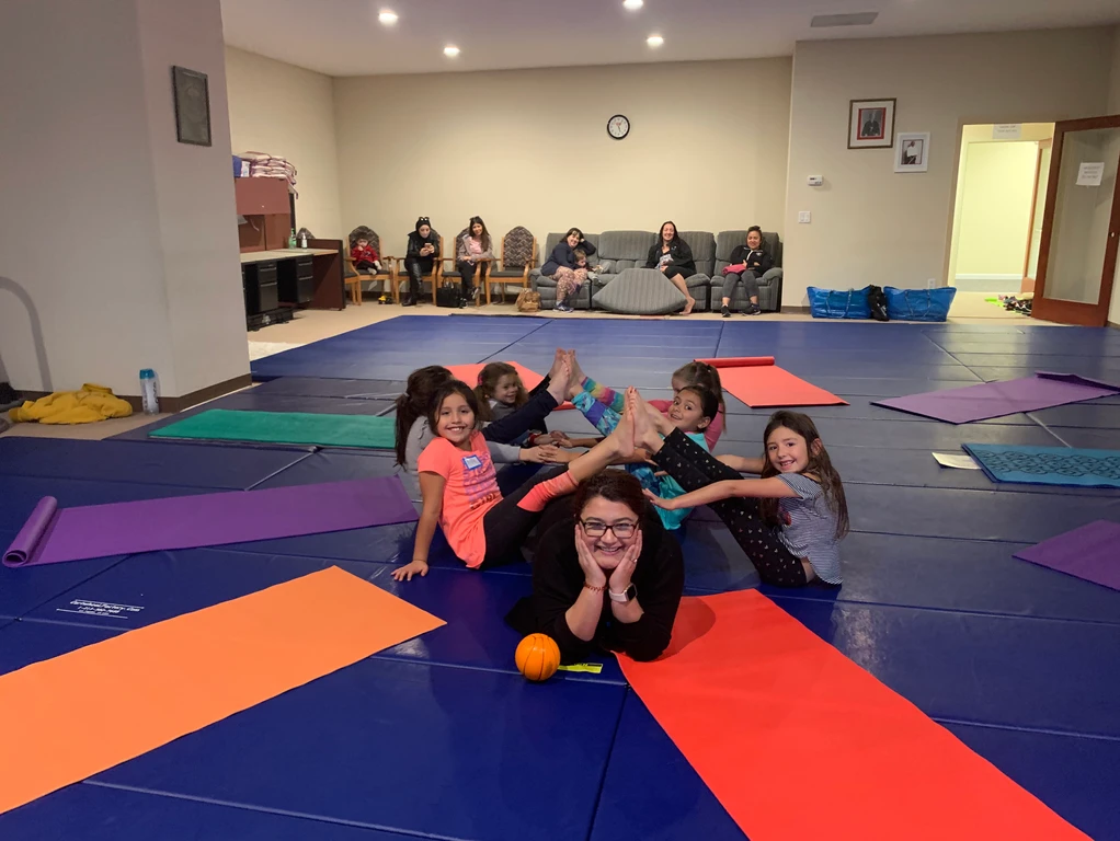 A group of children and an adult having fun on colorful mats in a room with observers in the background.