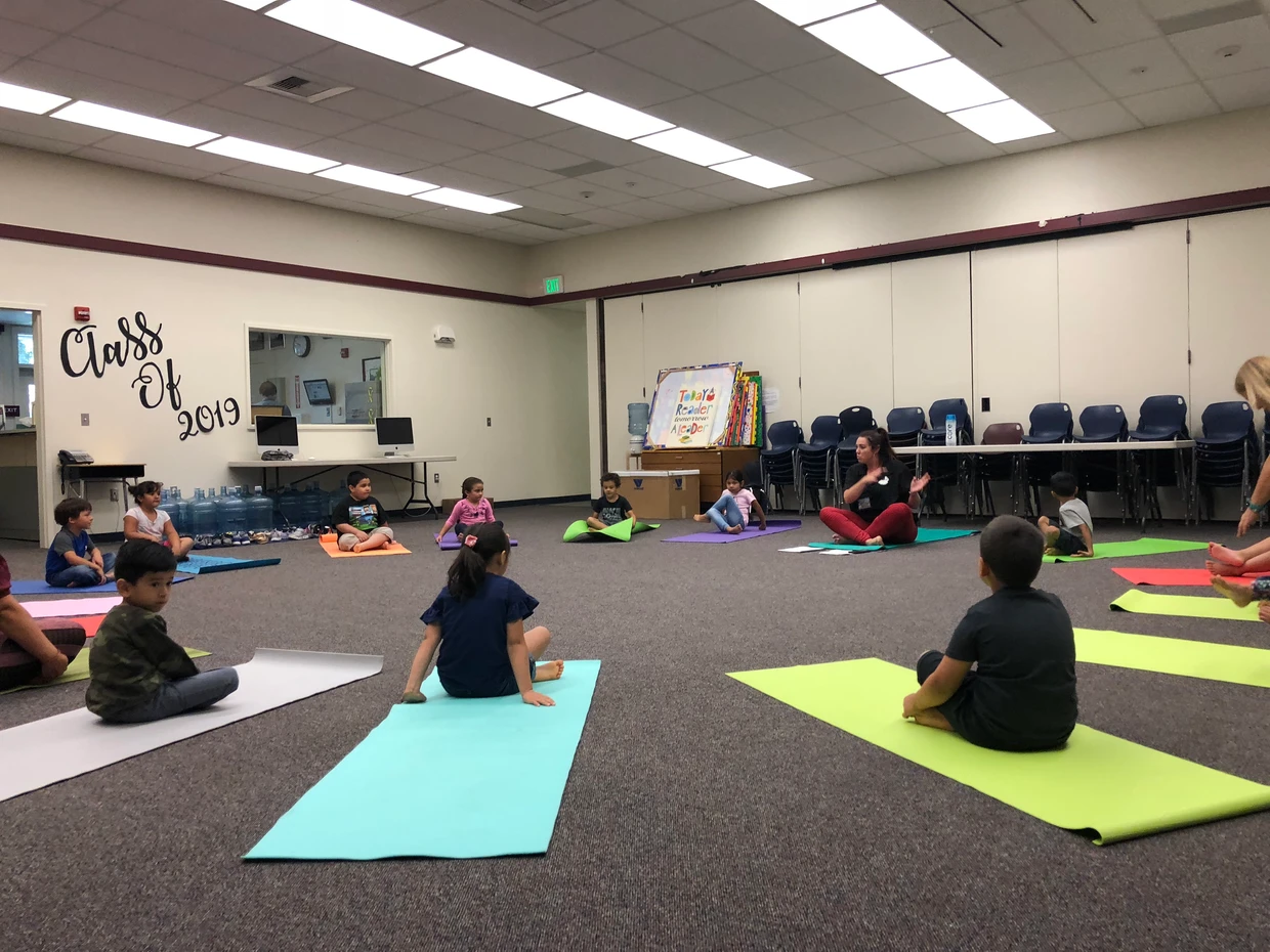 Children sitting on colorful mats in a classroom with an adult instructor, possibly during a group activity or class.