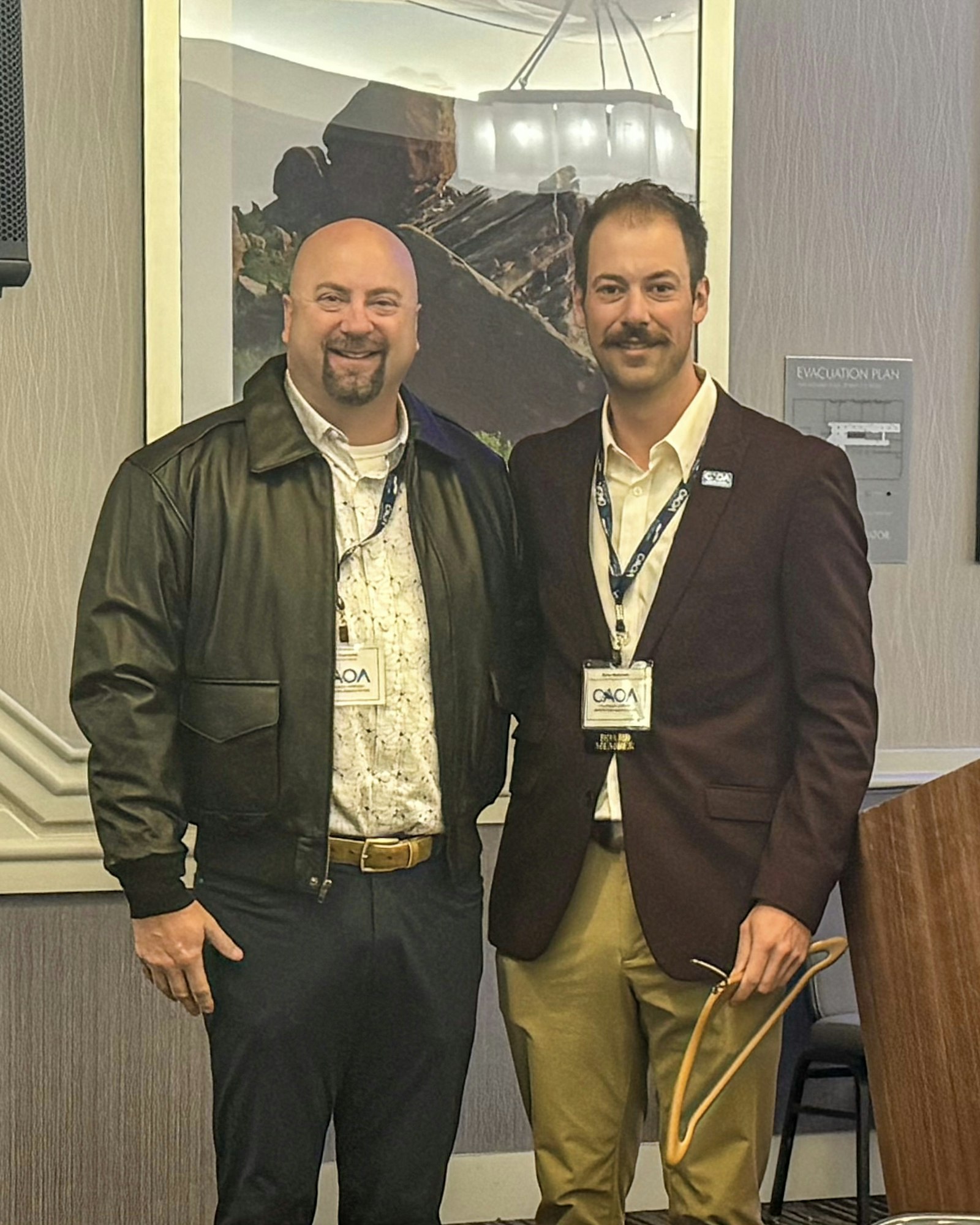 Two men are posing together, smiling, in a conference setting, wearing name tags and business attire. Background features a landscape photo.