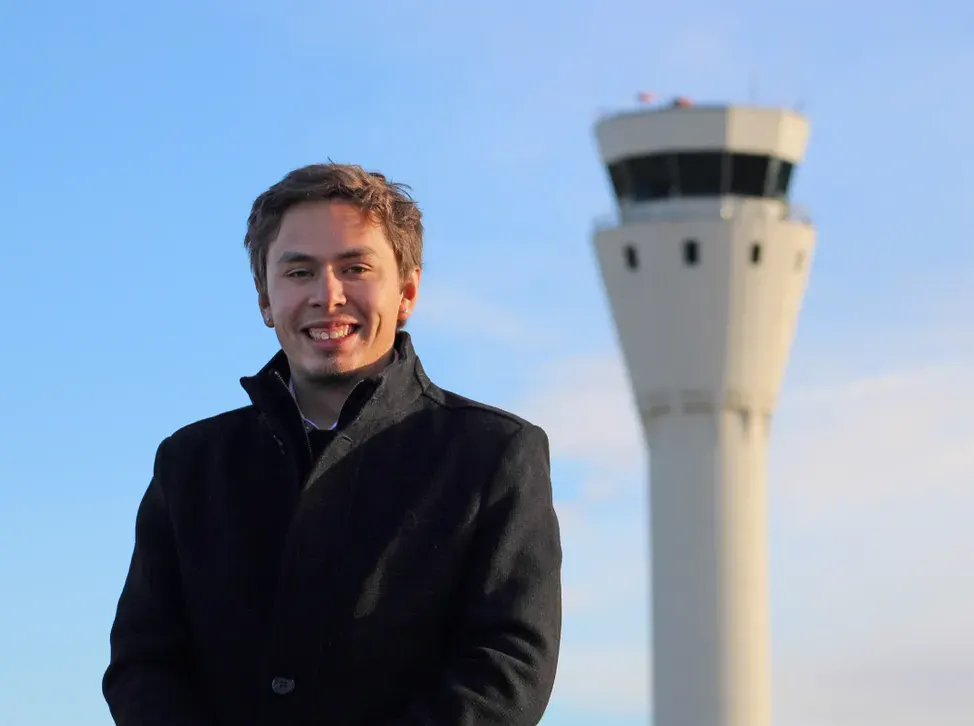 A smiling person in a coat standing in front of an airport control tower under a blue sky.