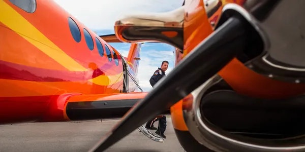A person stands near a vibrant orange aircraft, with a close-up view of the propeller and fuselage in the foreground.