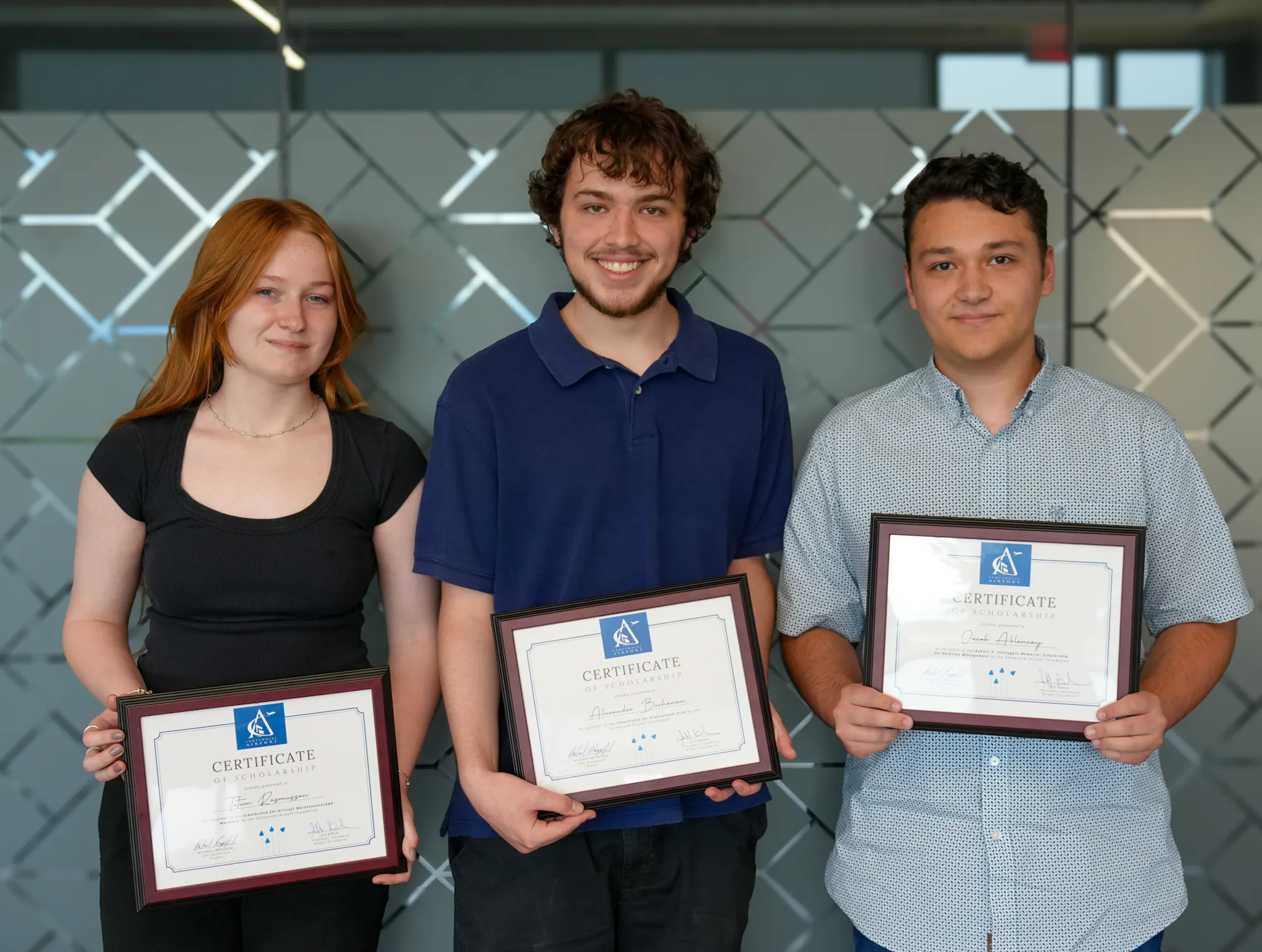 Three individuals hold certificates of scholarship, standing in front of a patterned glass wall, smiling for the photo.