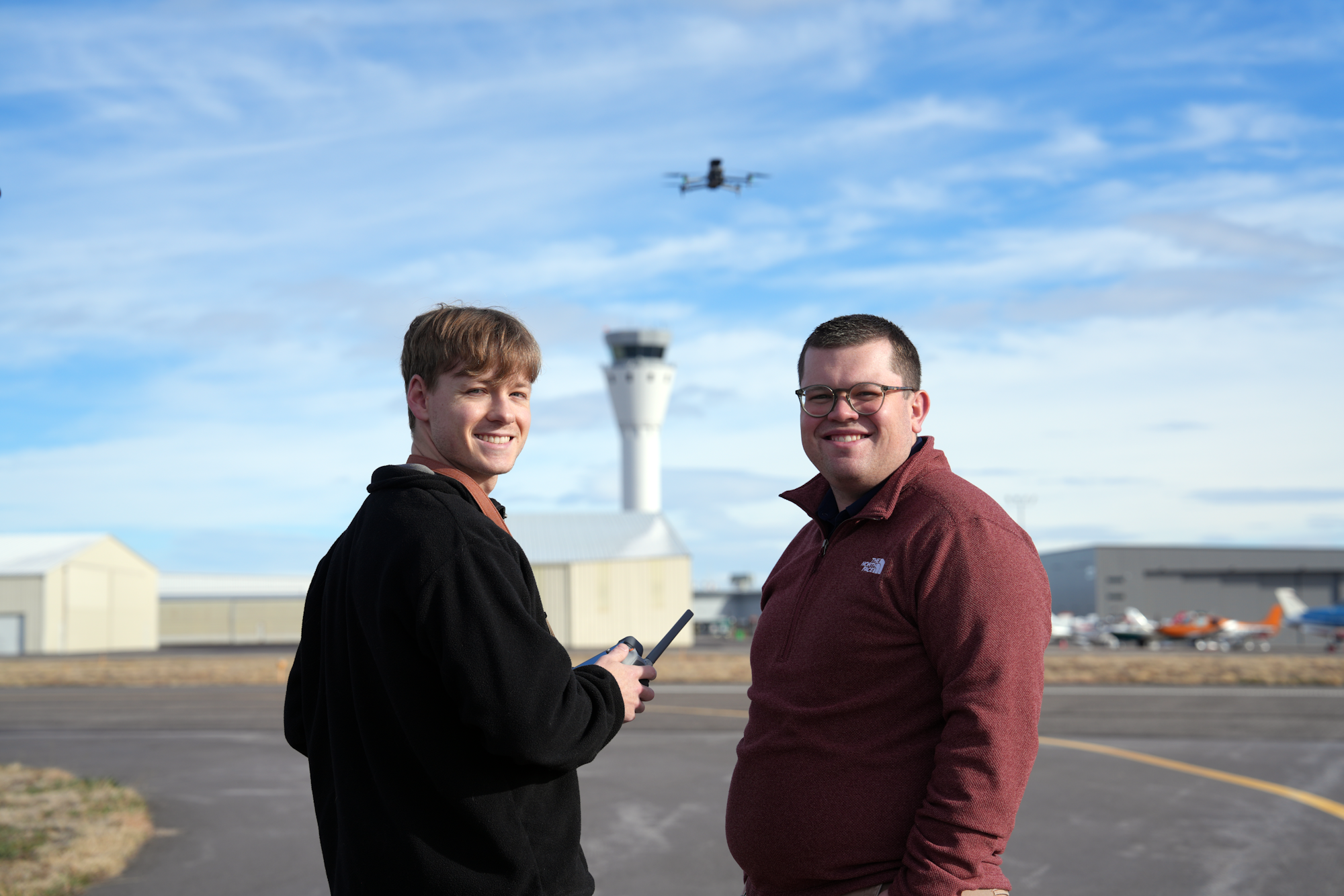 Two men are standing on an airport runway with a control tower and a drone flying in the background.