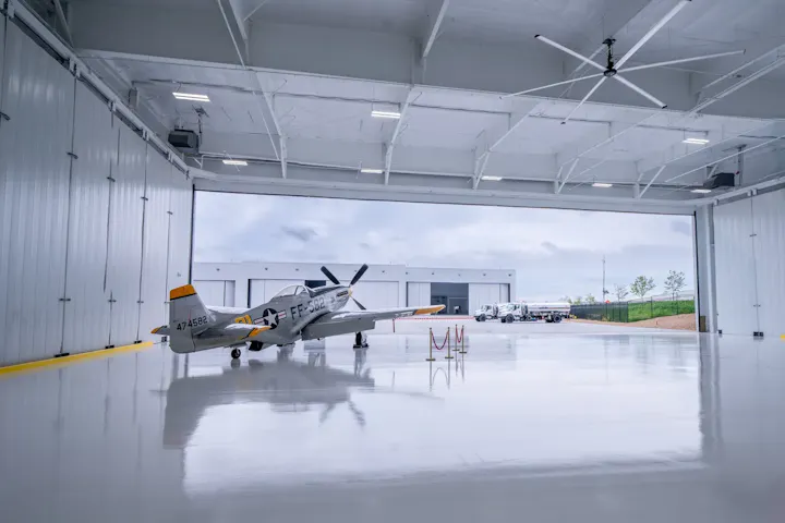 A vintage airplane parked inside a large, open hangar with a shiny floor, with trucks outside.