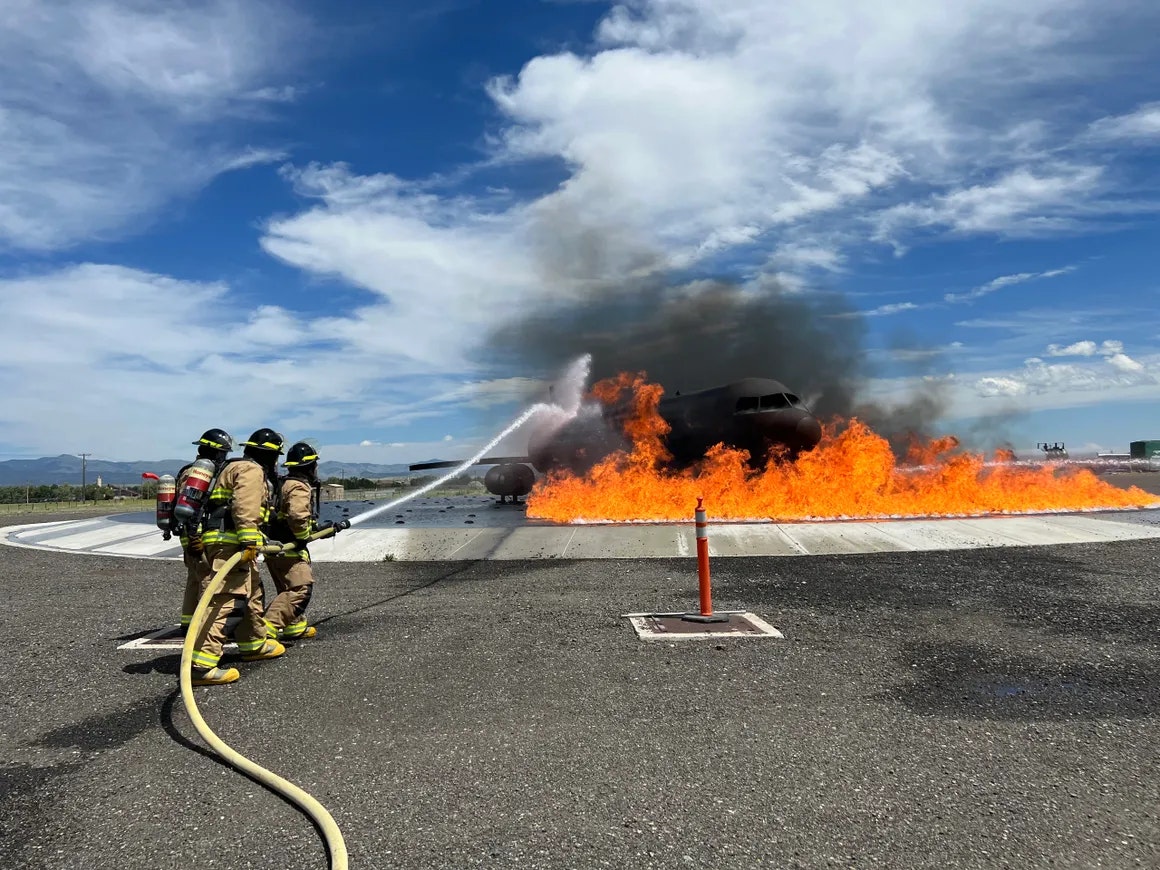 Firefighters extinguishing flames around a training aircraft with hoses.