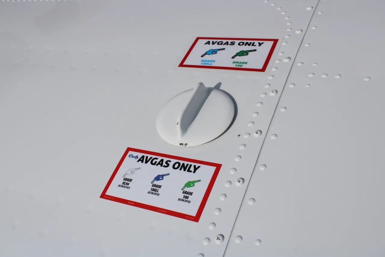 Airplane fuel cap with "Avgas Only" label showing compatible grades: 100LL, 100, UL94. Riveted surface.