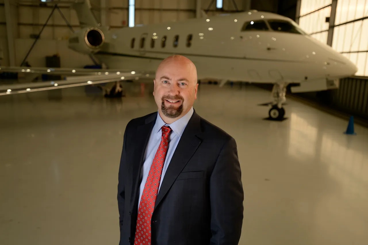 A man in a suit stands in a hangar with a private jet in the background.