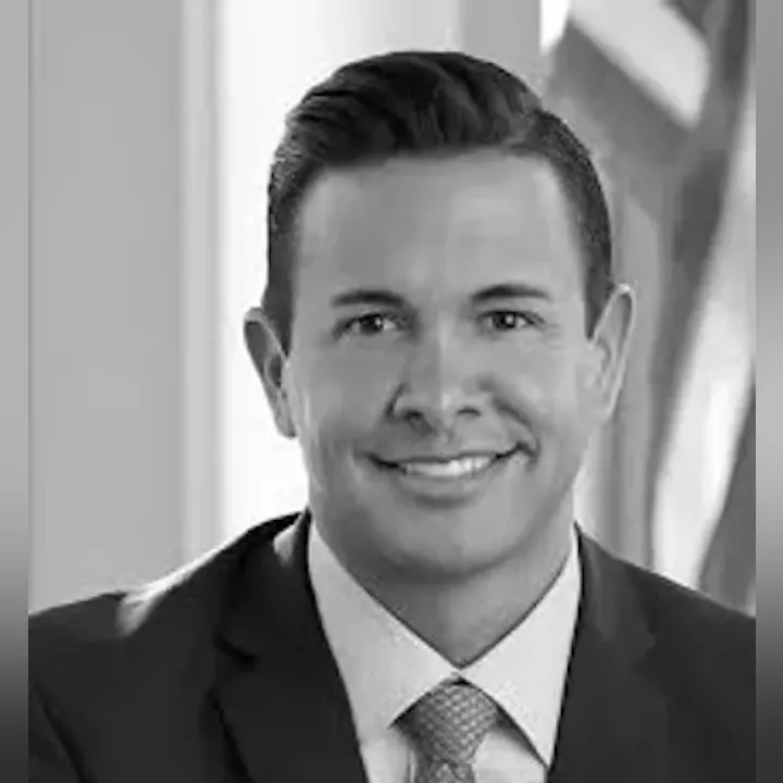 A grayscale portrait of a man in a suit and tie, smiling, with an American flag in the background.