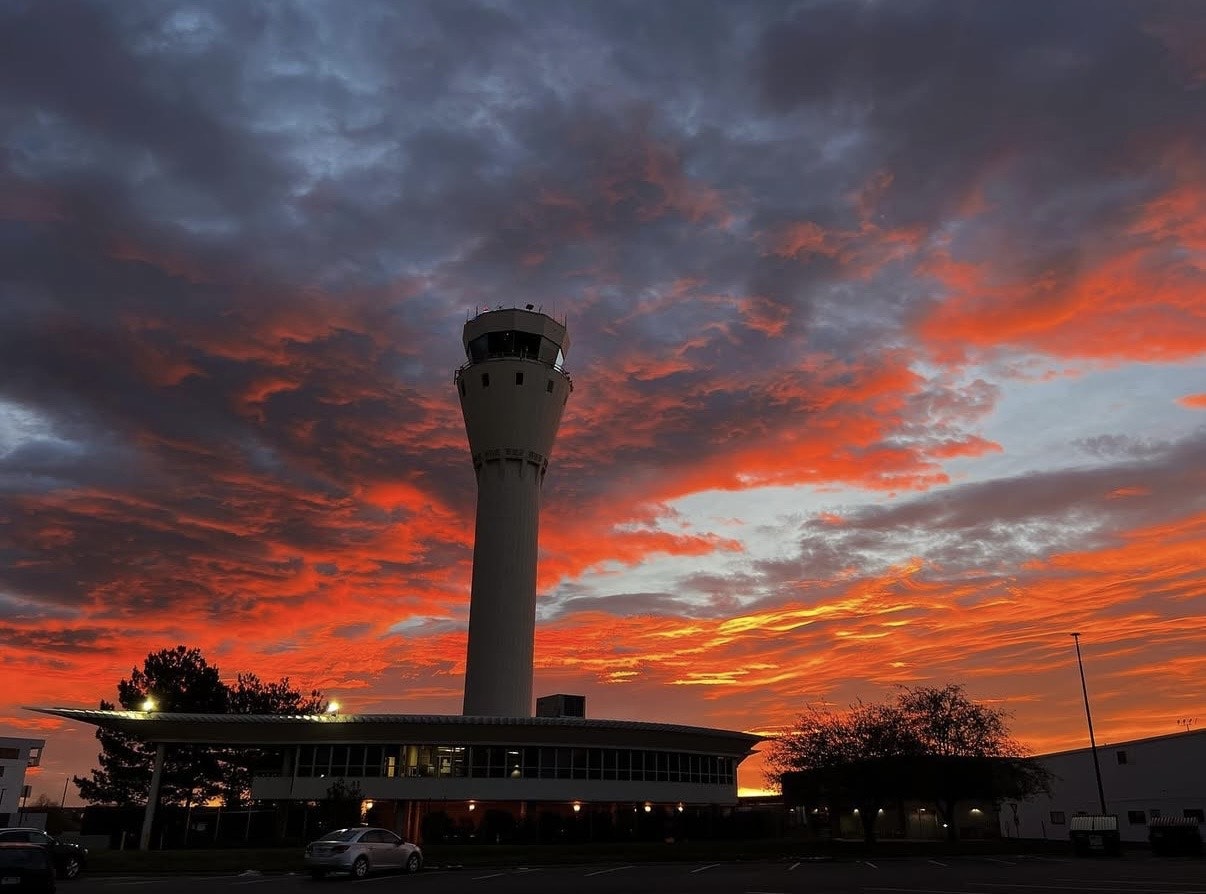 A dramatic sunset illuminates the sky with red and orange hues, silhouetting an airport control tower and buildings below.