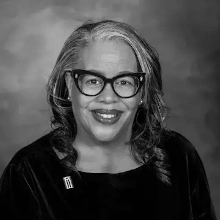 A person with glasses smiles, wearing a dark top. The photo is black and white.