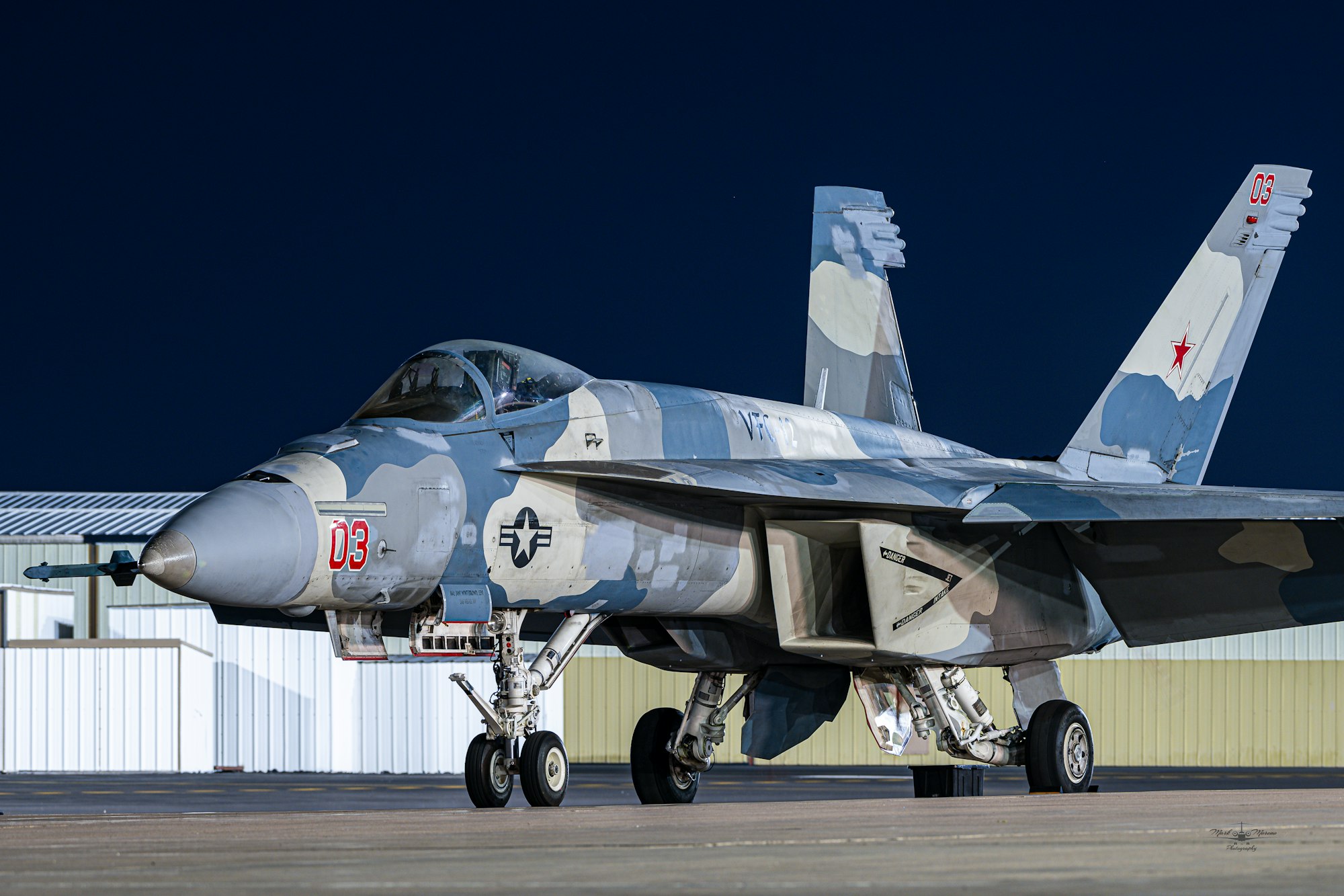 The image features a military fighter jet with a unique camouflage pattern, parked on a runway at night.