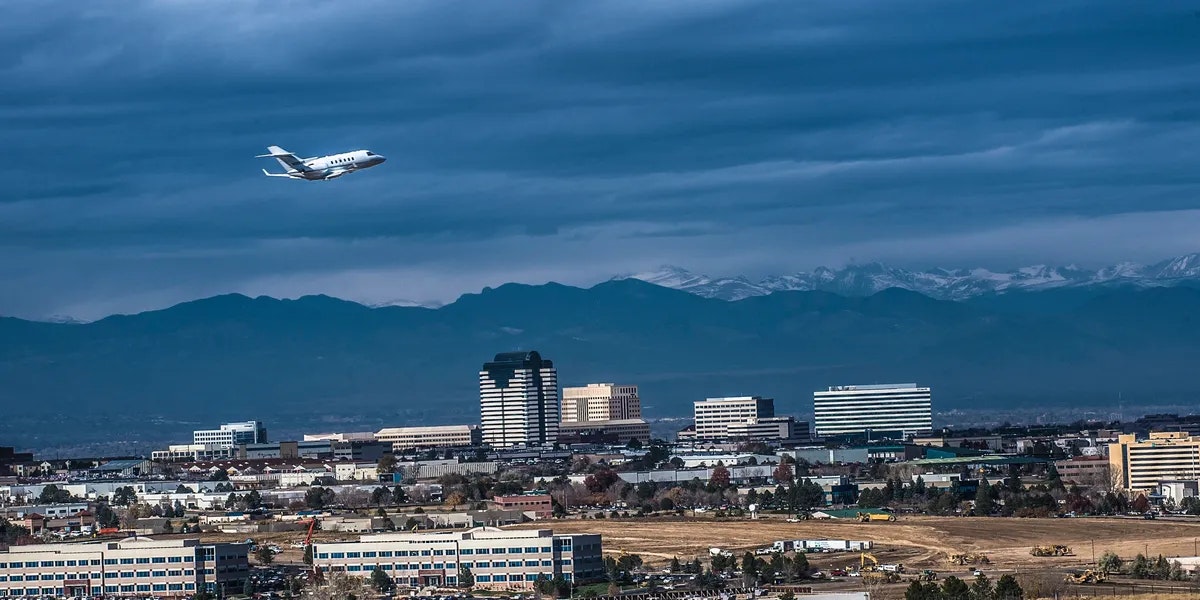 Airplane flying over a cityscape with tall buildings and mountains in the background under a cloudy sky.