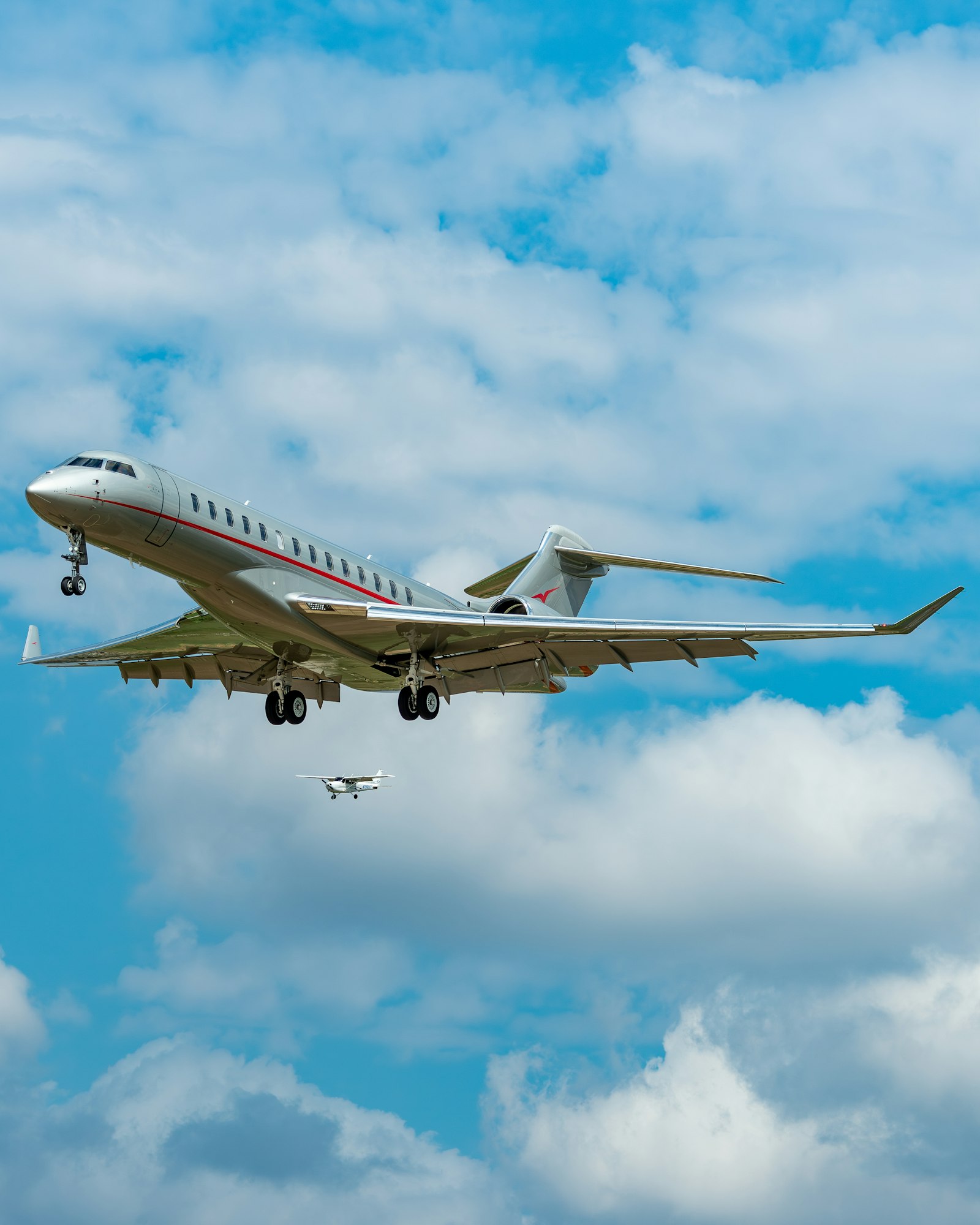 A large private jet descends through a cloudy sky, with a smaller aircraft flying below it.