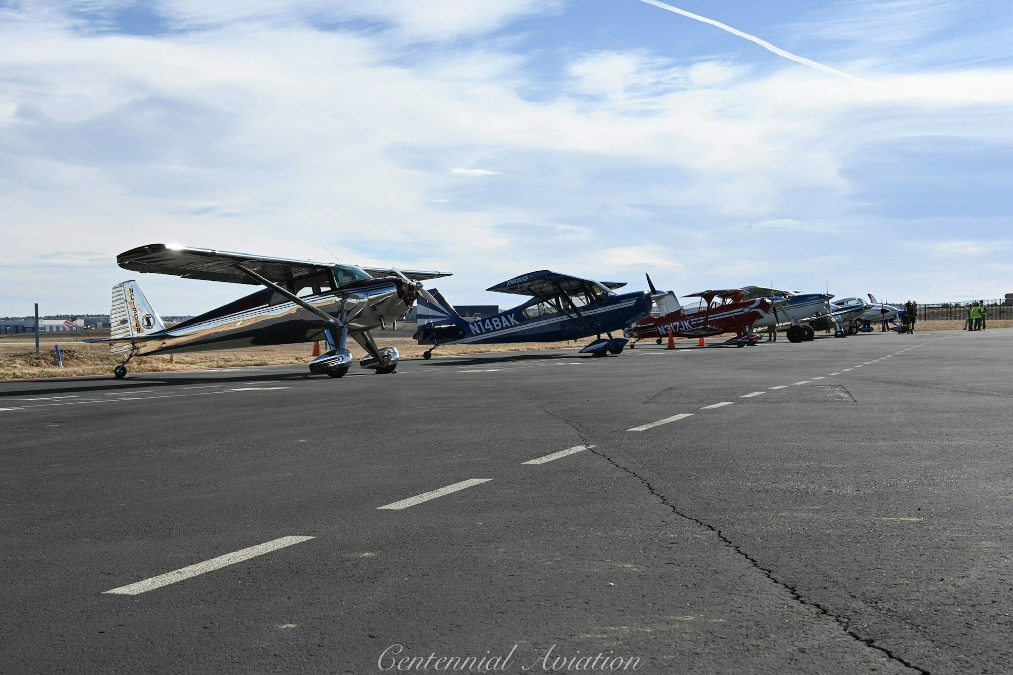 Several small aircraft are lined up on a runway, with blue skies and distant scenery visible.