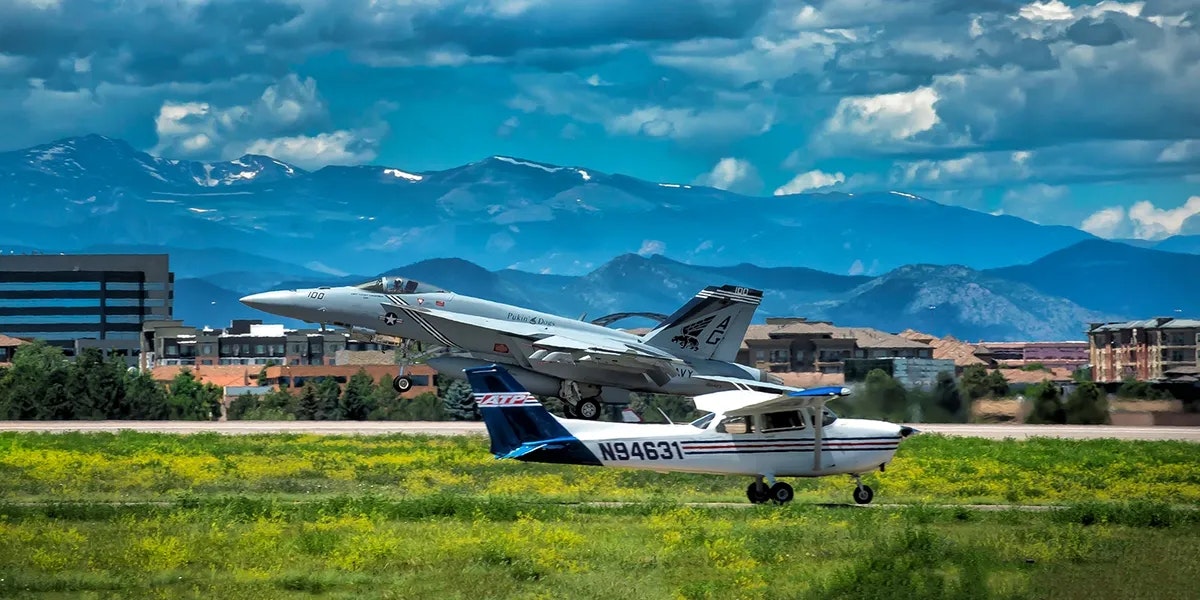 A fighter jet takes off behind a small propeller airplane on a runway with mountains and buildings in the background.