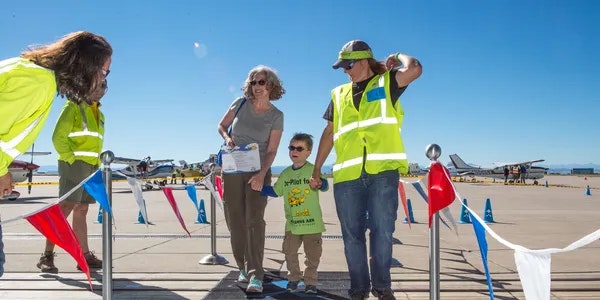 A woman and a child are greeted by staff in safety vests at an airfield with planes in the background, likely at an event.