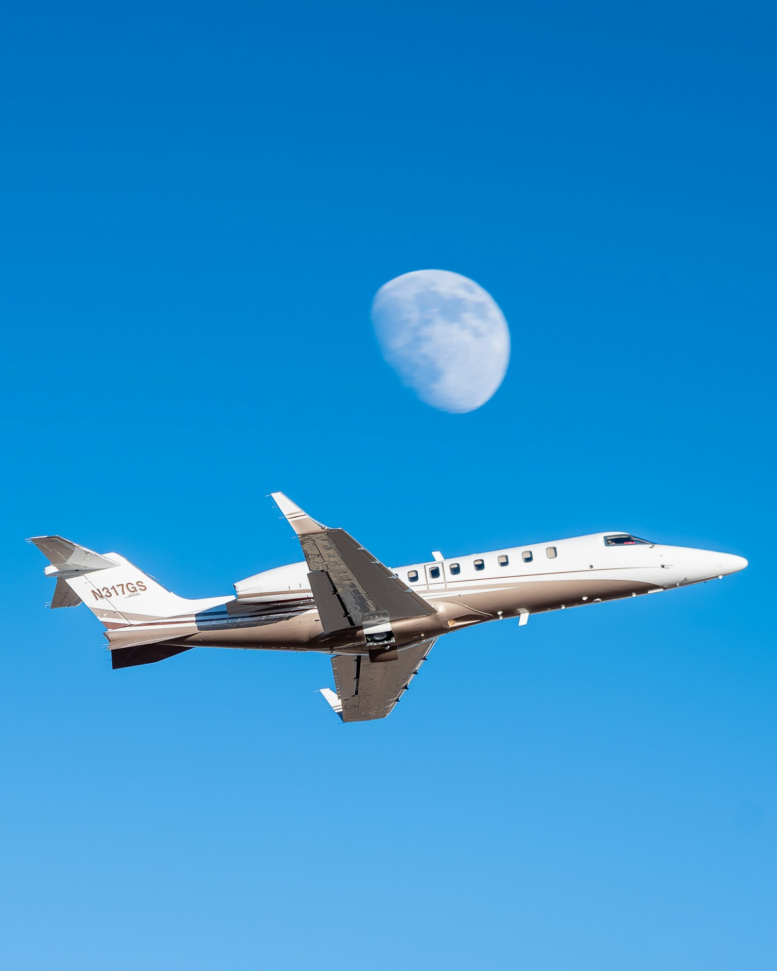 A jet aircraft flying against a clear blue sky, with the moon partially visible in the background.