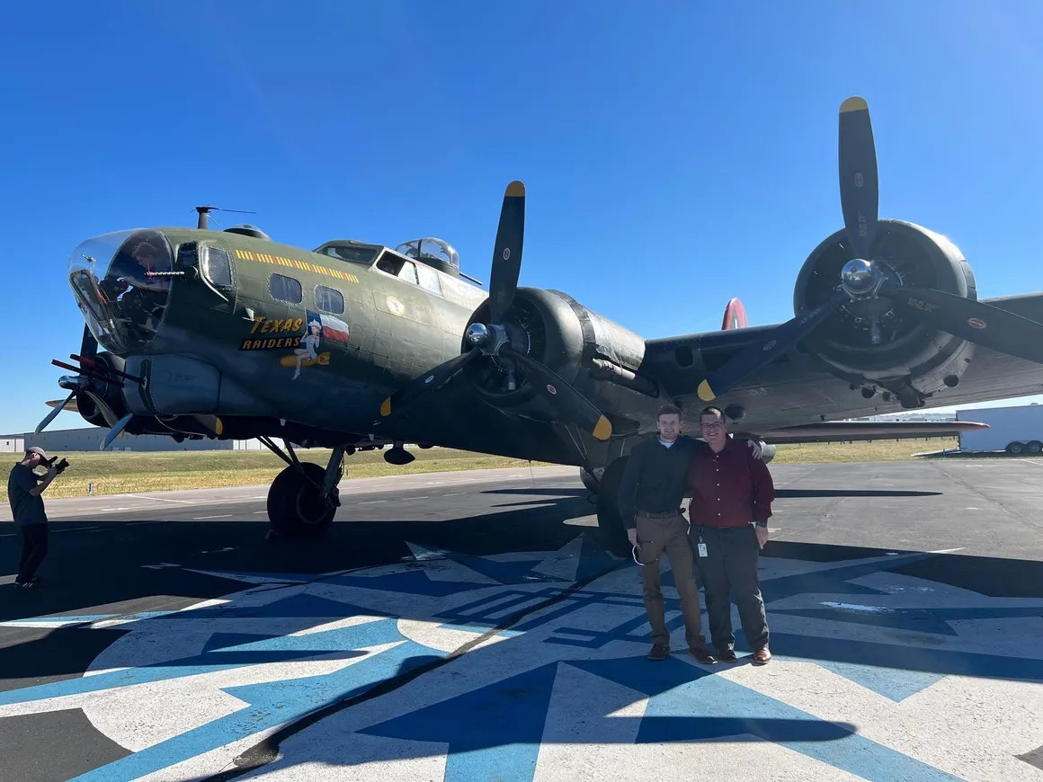 Two people standing in front of a vintage military aircraft with a "Texas Raiders" insignia, another person taking a photo.