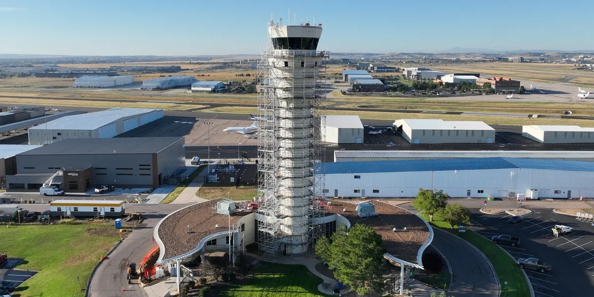 Airport control tower surrounded by scaffolding, with hangars and small aircraft in the background.