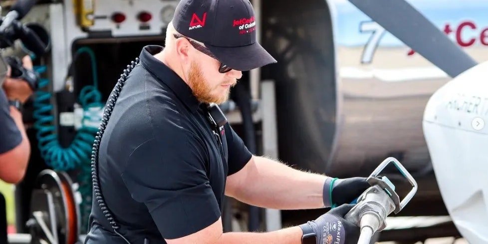 A person refuels an aircraft, wearing gloves and a cap, near a fuel truck.