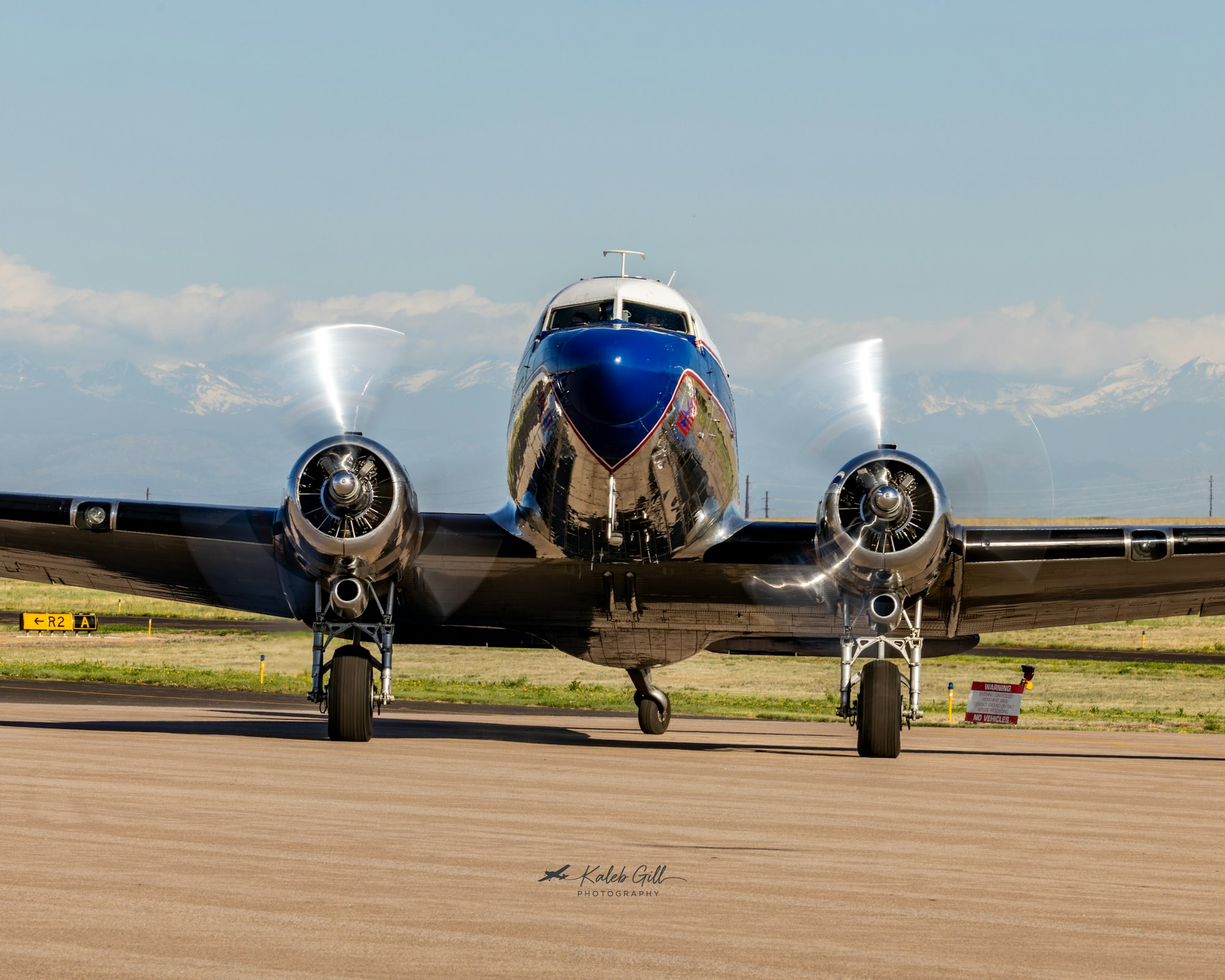 A classic aircraft with spinning propellers, positioned on a runway with mountains in the background under clear skies.