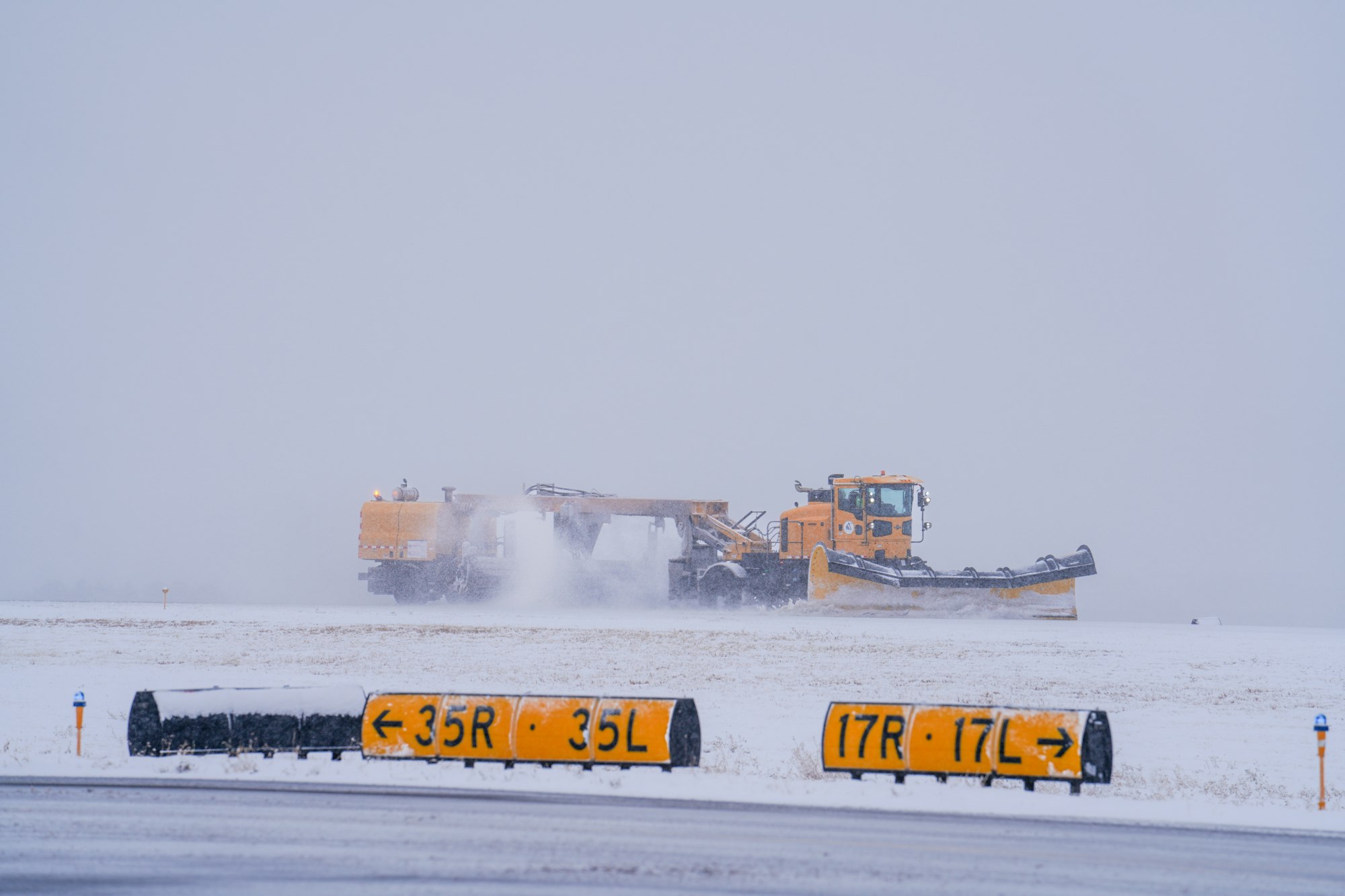 A snow plow clears snow on an airport runway during a snowstorm, with runway markers visible in the foreground.
