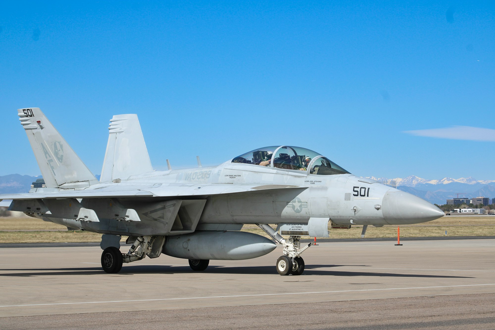 A military jet, likely a fighter aircraft, is on a runway with mountains in the background and a clear blue sky.