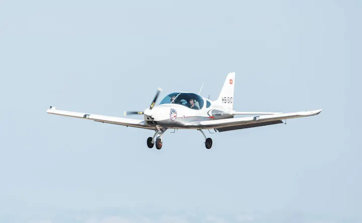 A small white aircraft with a visible pilot is in flight against a clear sky.