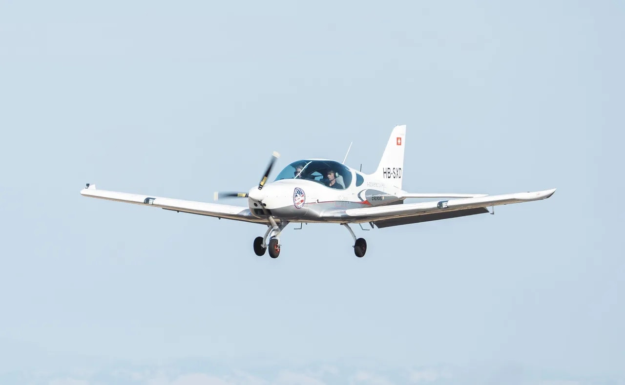 A small white aircraft with a visible pilot is in flight against a clear sky.