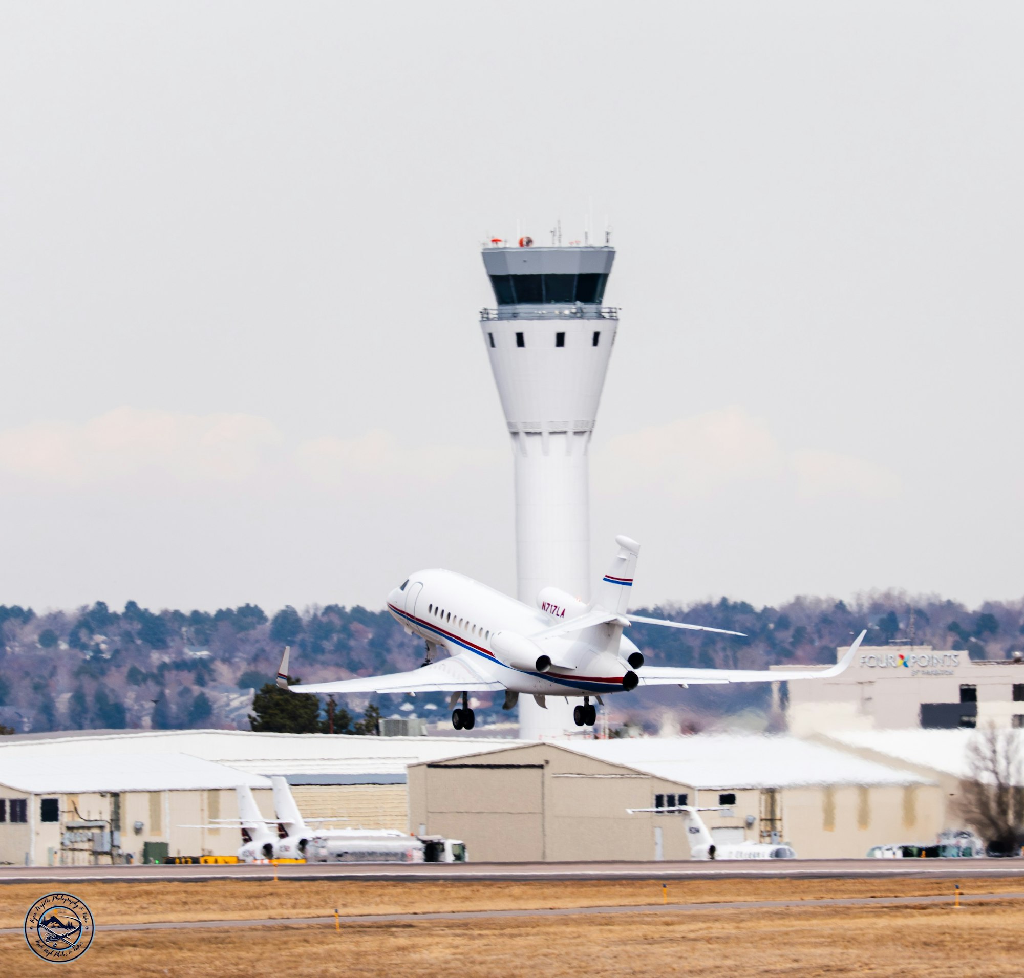 A jet airplane departing an airport with a control tower and hangars in the background on a clear day.