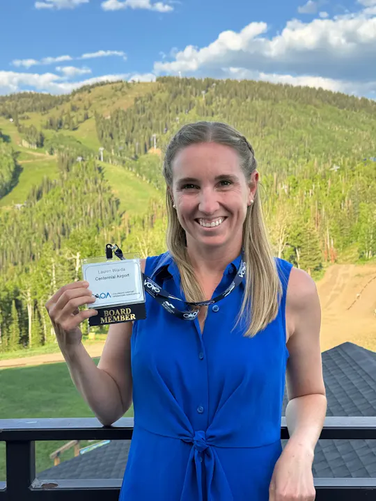 A person in a blue dress holds a "Board Member" badge in front of a scenic, mountainous background.