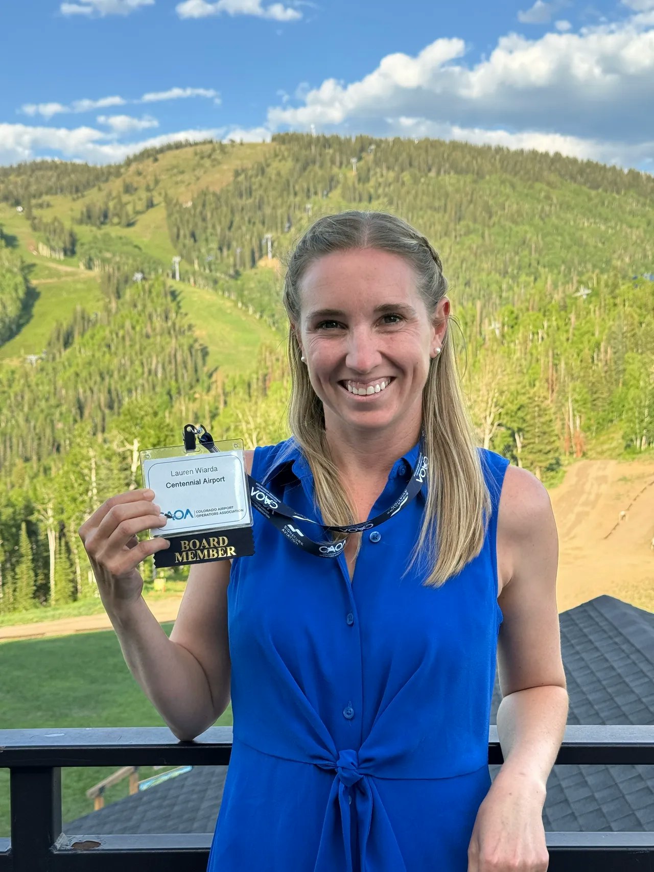 A person in a blue dress holds a "Board Member" badge in front of a scenic, mountainous background.