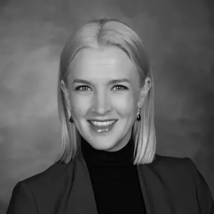 A black and white portrait of a smiling person with straight hair, wearing a jacket and earrings, against a neutral background.