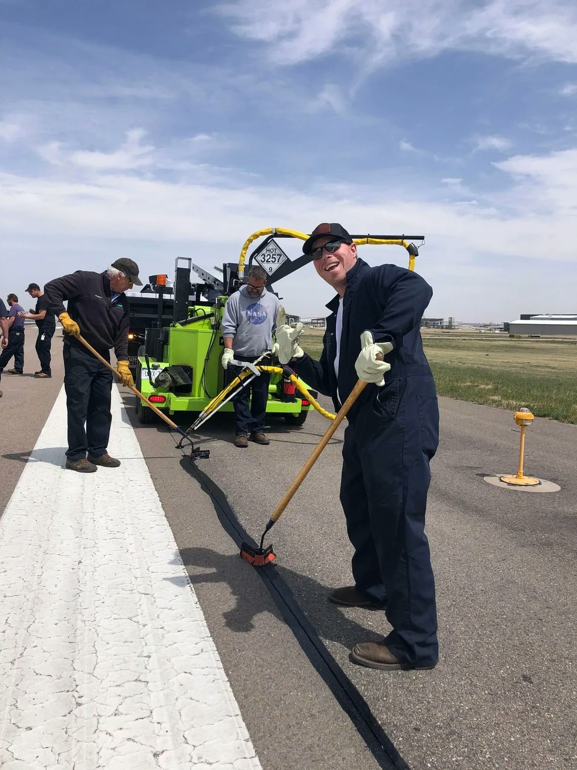 People working on a runway, sealing cracks with tar using brooms and a machine.