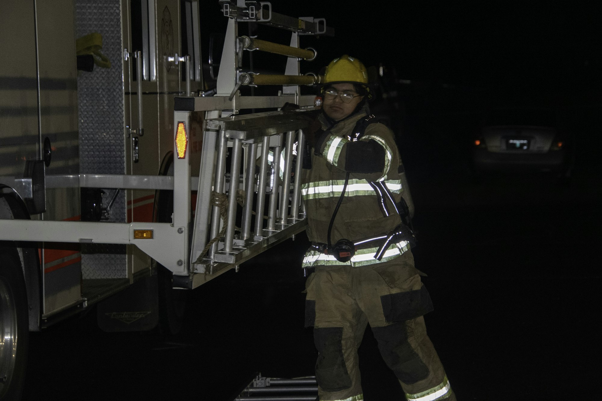 A firefighter in gear is handling a ladder at night, near a fire truck. Safety equipment is visible on their uniform.