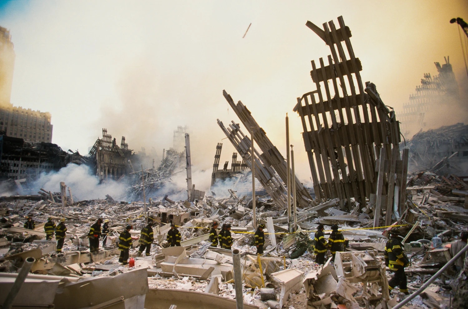 Firefighters amid rubble and smoke at a disaster site with twisted metal structures.