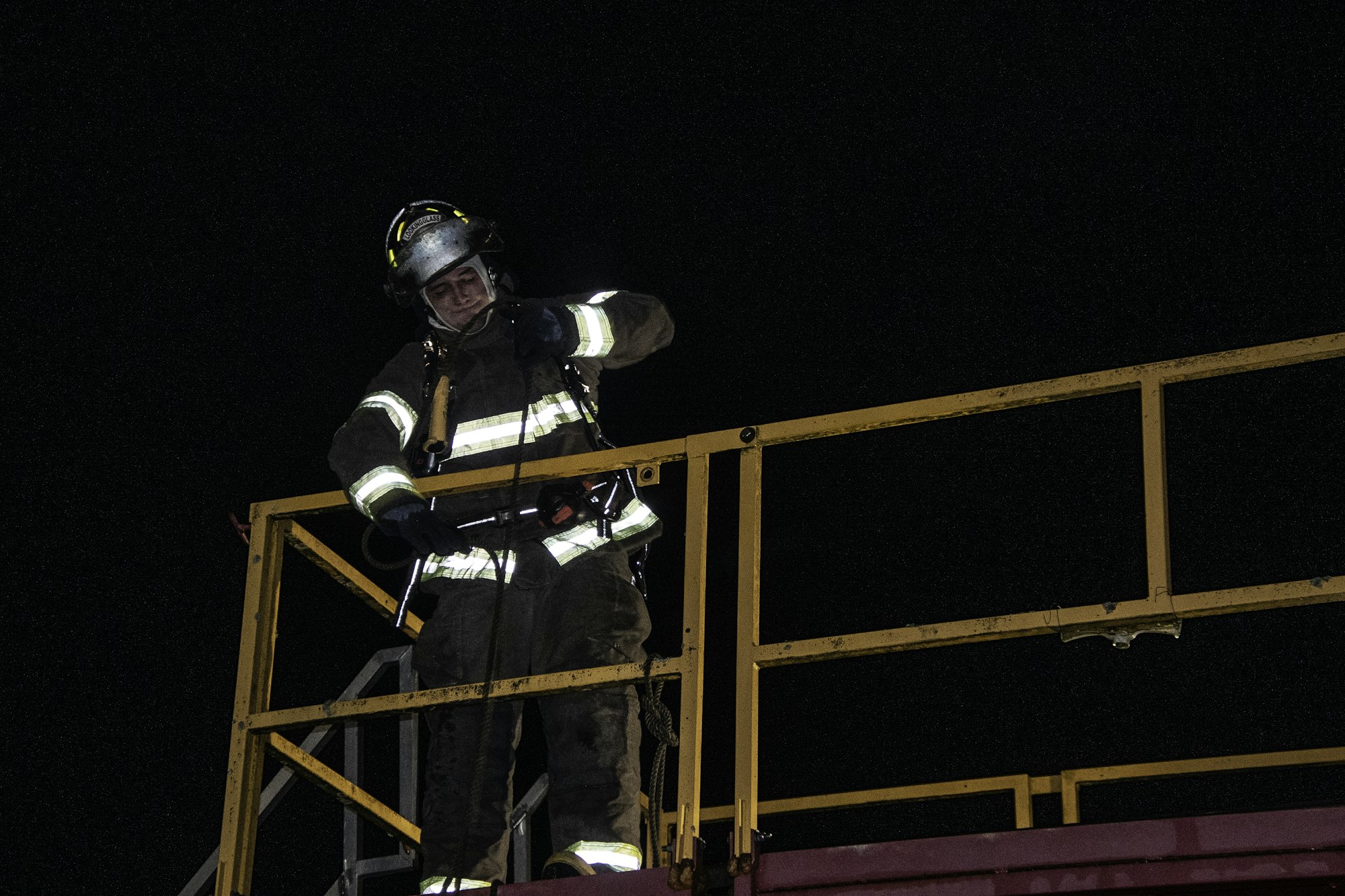 A firefighter in protective gear stands on a platform at night, preparing equipment or assessing the situation.