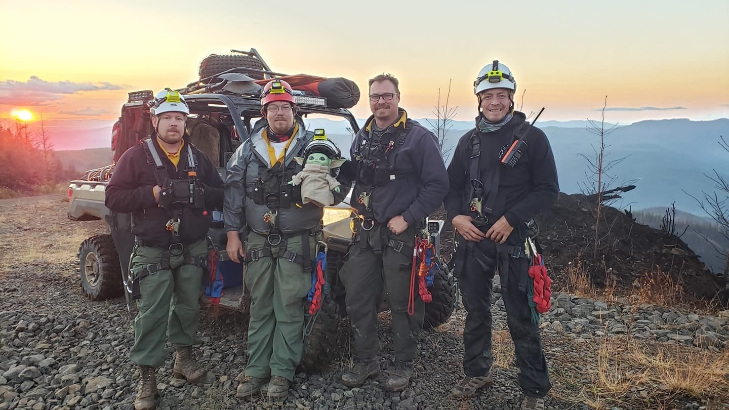 Four people in safety gear with an off-road vehicle and a plush of an iconic character, set against a scenic sunset backdrop.