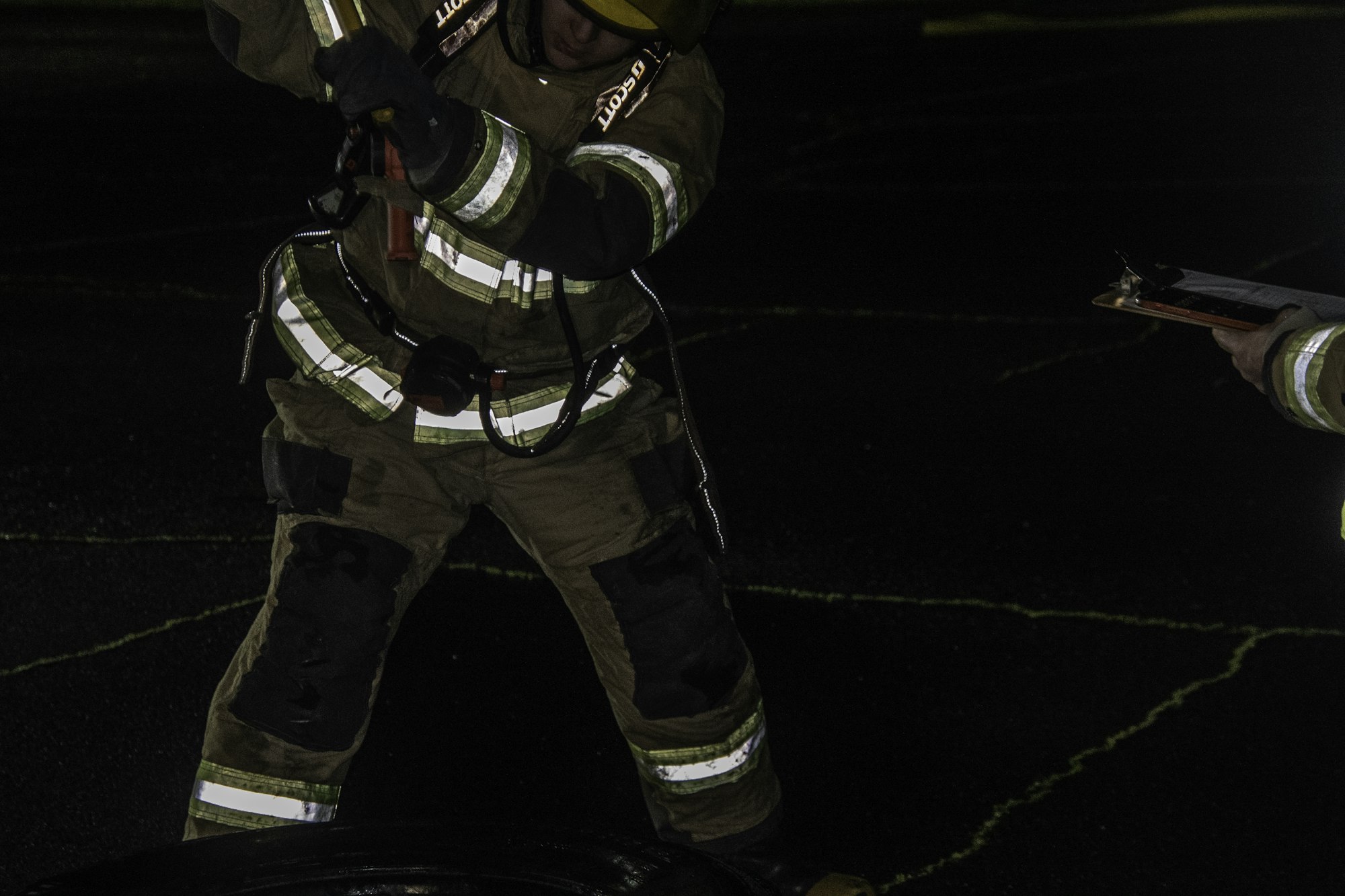 A firefighter is shown using a tool while another person holds a clipboard, likely during a night training exercise.