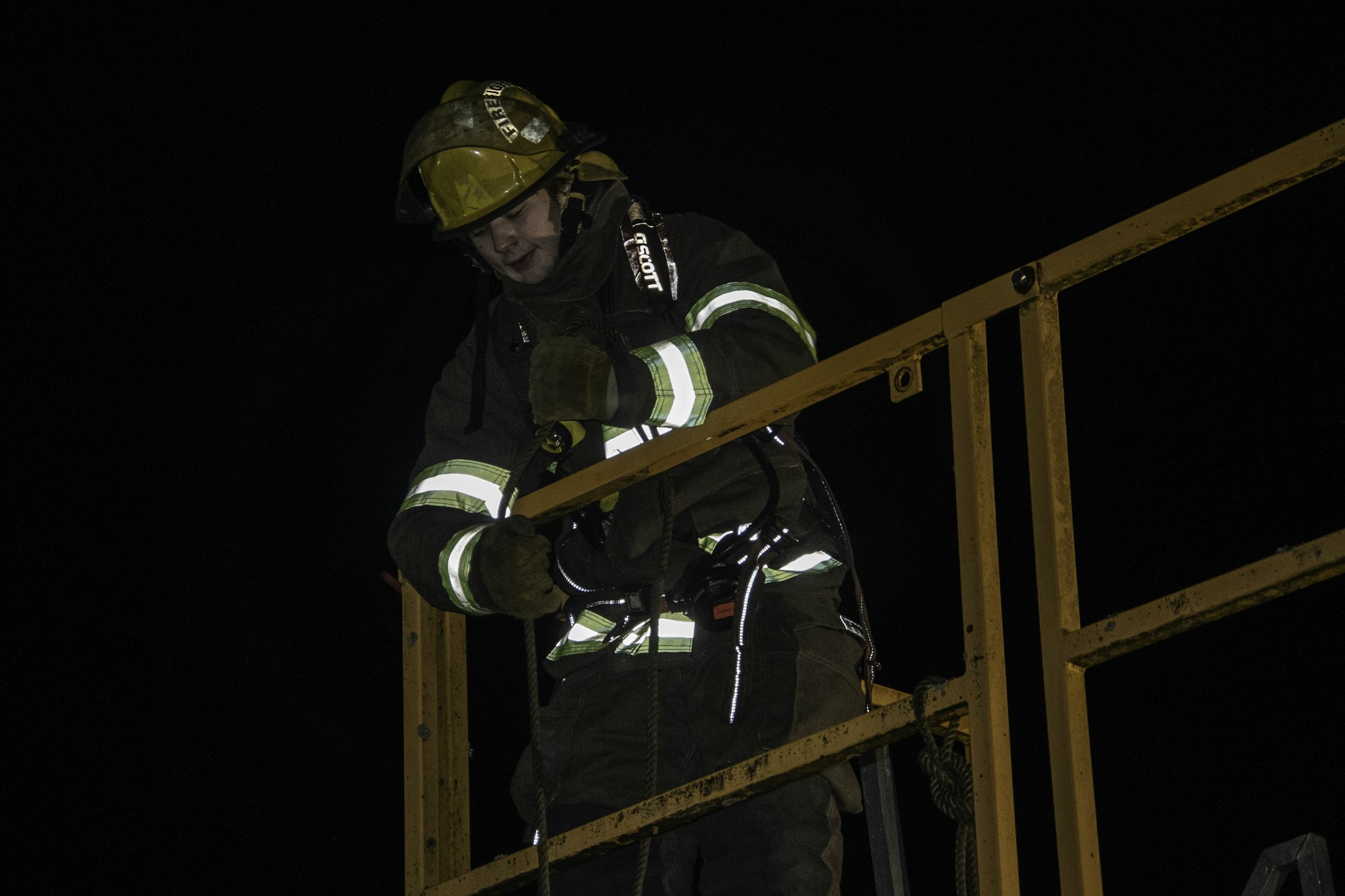 A firefighter in gear working on a ladder at night, focused on a task with safety equipment in use.