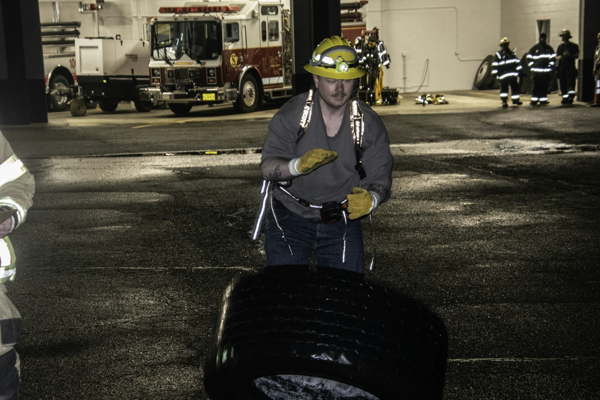 A person wearing a helmet and gloves appears to be working with a tire inside a fire station, with fire trucks in the background.